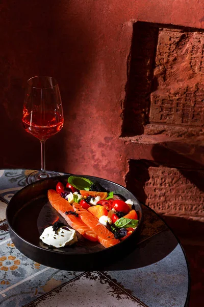 Salad with salmon fillet, tomatoes, basil, feta cheese, caviar and sauce in a plate on the table with a glass of wine against a brown wall