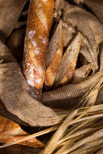 Whole French baguette, next to it are sliced pieces of bread, in a linen sack of flour. The bag stands on a wooden background. In the background a cut baguette and ears of wheat.