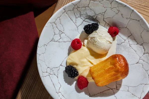 Savarin soaked in rum with vanilla ice cream and vanilla cream. Topped with raspberries and blackberries. The dessert lies in a round, deep plate with a pattern in the form of cracks. The plate stands on a wooden table with a traditional oriental blu