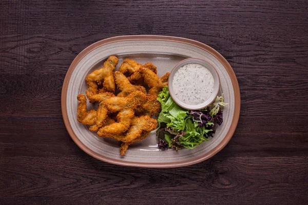 Fried frog legs with salad, arugula and parsley sauce on an oval plate and on a wooden background. Horizontal orientation