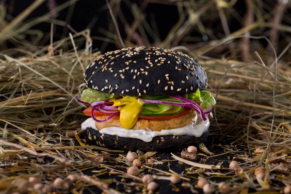 Burger with black sesame bun, cutlet, yellow sauce, salad, fresh onions, cucumbers and mayonnaise on the table with chickpeas and hay sprinkled on a black background. Horizontal orientation