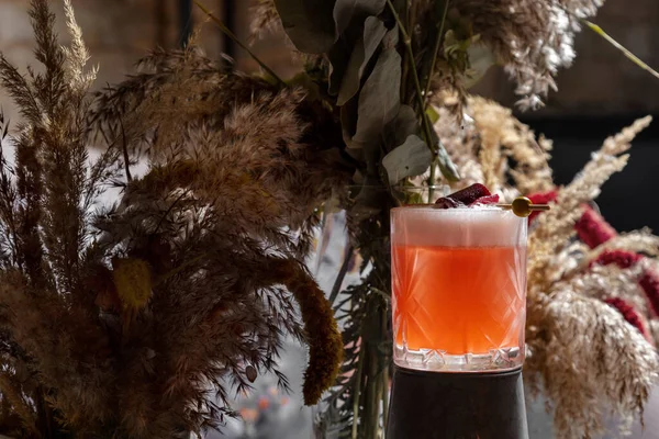 Cocktail with marmalade and olive on a stick in a glass that stands on a stand against a background of dried flowers