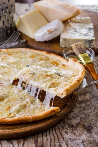 Pizza with different types of cheese on a wooden table, a piece lifted with a spatula and lying on the table with a grater, cutlery and four types of cheese lying aside on a wooden board