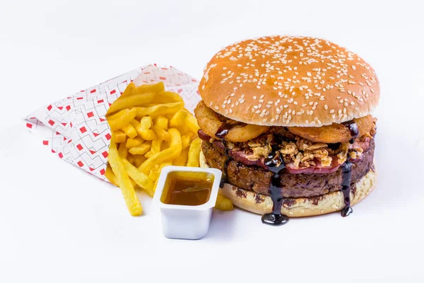 Burger with sesame bun under dark sauce with cutlet, ham, onion chips and onion rings with fries and sauce on a white background. Horizontal orientation