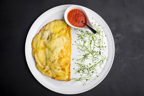 Protein omelet with mushrooms on a white, round plate. The plate is strewn with microgreen onion sprouts. On the plate is a white gravy boat with tomato sauce and a spoon inside. The plate stands on a black background.