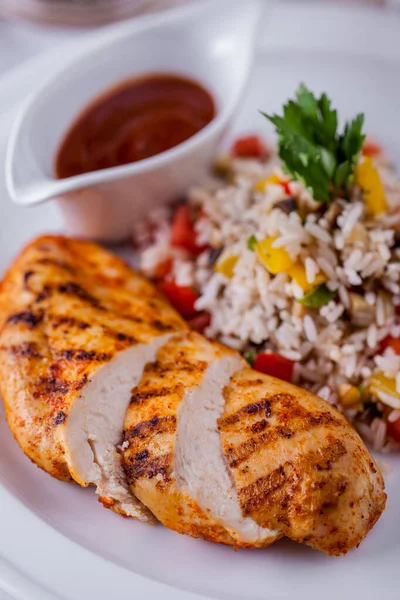 Grilled chicken fillet is sliced in portions, next to boiled rice with stewed vegetables, food is on a white plate, next to it is a sauceboat with tomato sauce.