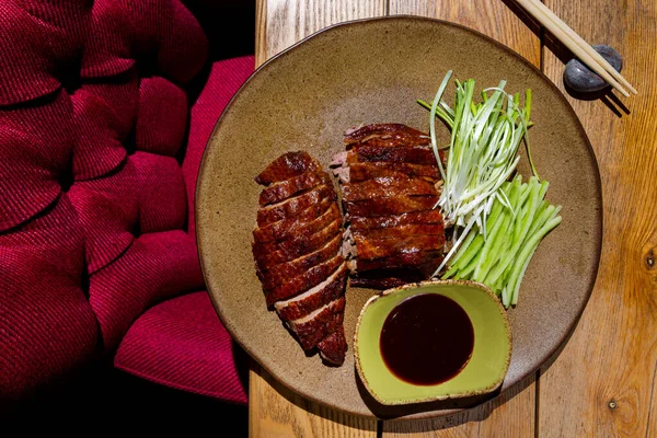 Fried chicken breast with leeks and cucumber in a plate with sauce in a bowl and chopsticks lying on the table and a red chair on the floor