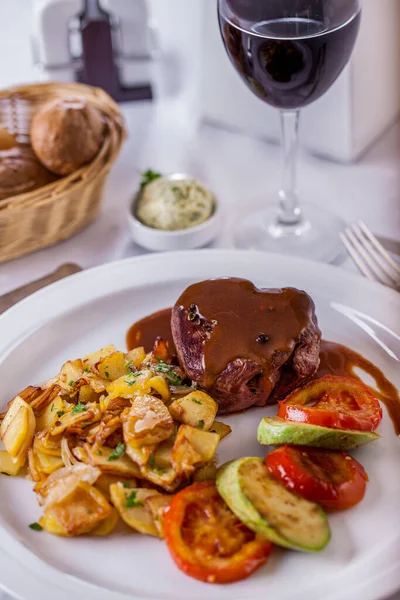 Grilled meat steak with sauce with potatoes, tomatoes, parsley on a round plate with a basket of bread and a glass of wine on a light background. Vertical orientation