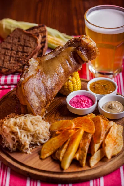 Bir bardak birayla domuz budu, fırında patates, tost, mısır, sos, lahana, ahşap bir masanın üzerinde beyaz çizgili kırmızı bir masa örtüsü ve kırmızı ve beyaz kareli bir peçete.