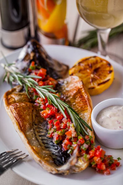 Grilled stuffed mackerel with lemon, rosemary, sauce in a gravy boat on an oval white plate and a glass of white wine on a wooden table. Vertical orientation 