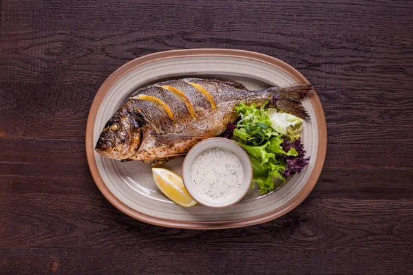 Grilled dorado stuffed with lemon slices with white sauce and arugula salad on an oval plate and a wooden table. Horizontal orientation 