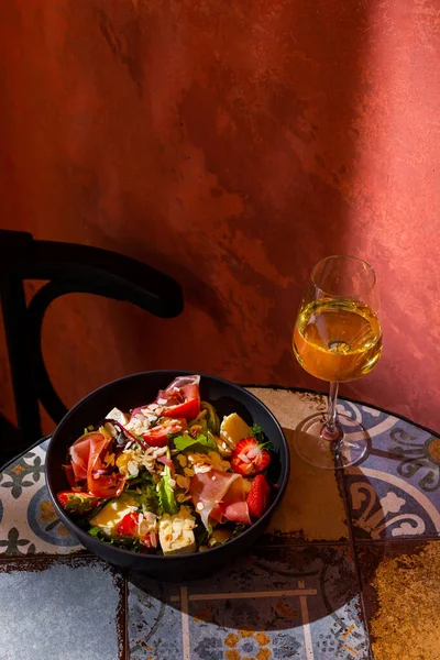 Salad with prosciutto, strawberries, mango, cheese, almond slices, arugula, salad and sauce in a plate on the table with a glass of wine against a brown wall
