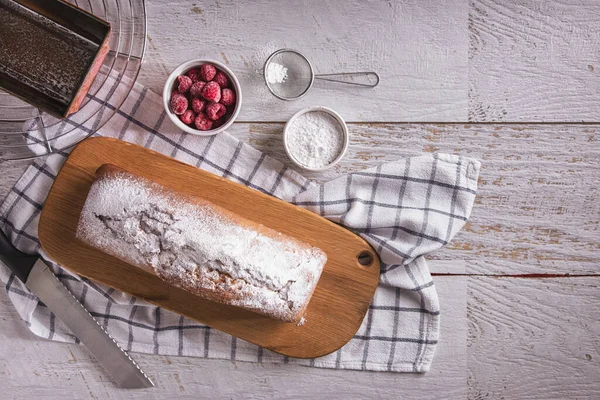 Sweet white bread with powdered sugar and raspberries lying on a wooden board with a towel and a knife on the table
