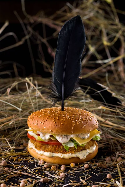 Burger with sesame bun and black feather stuck on top, cutlet, white sauce with parsley and mustard, lettuce, cheese and tomatoes on the table with chickpeas and hay on a black background. Vertical orientation