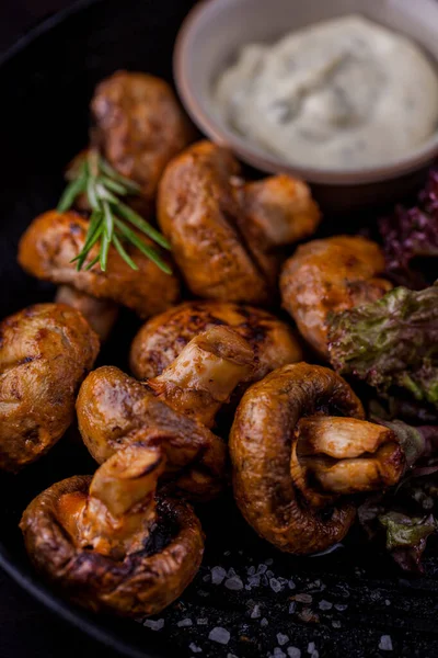 Biberiye filizli ızgara şampiyon mantarları, kaba tuz ve marul. Mantarların yanında seramik sos kabı var. İçinde sarımsaklı mayonez var. Siyah, metal kızartma tavası.