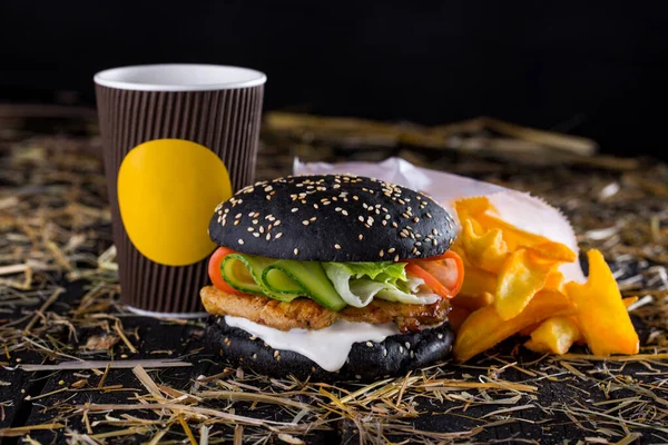 Burger with sesame black bun, cutlet, pickled cucumbers, lettuce, tomatoes and white sauce on the table with chickpeas, fries, a glass and a sprinkle of hay on a black background. Horizontal orientation