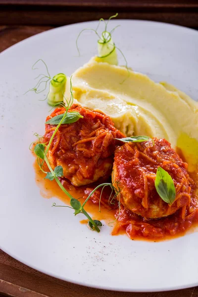 Fish cakes in tomato sauce, basil leaves and carrots with mashed potatoes and microgreen pea sprouts. Zucchini rolls are next to the mashed potatoes. The food is on a white, round plate. The plate stands on a wooden background.