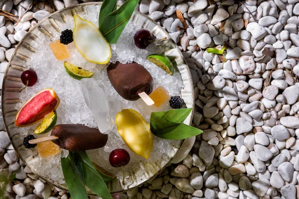 Chocolate ice cream on a stick, cherry, lime, sorbet in lemon peels, blackberries, banana leaves and marmalade lying in ice plates on stones
