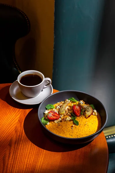 Pumpkin porridge with strawberries, apple, mint, avocado, sesame seeds, cheese and walnuts in a plate on the table with a cup of coffee