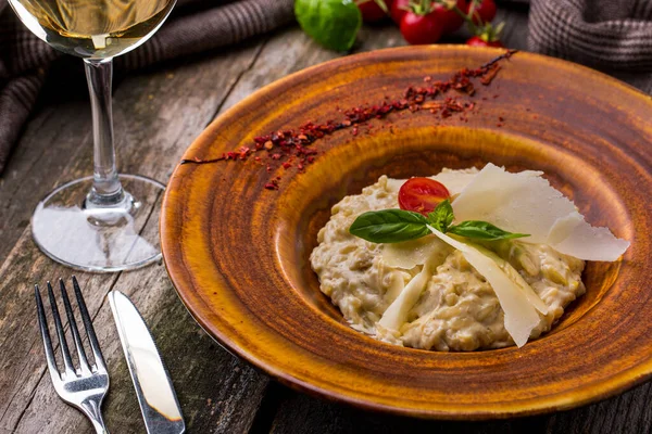 Risotto with porcini mushrooms, onions and cheese sauce. On top are Parmesan slices, a basil leaf and half a cherry tomato. The food is in a round, brown, ceramic plate with paprika sprinkled on board. The plate stands on an oak, wooden background. N