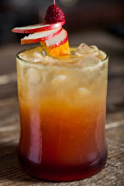 A cocktail with ice, apples, orange peel and raspberries on a skewer in a glass that stands on a wooden table