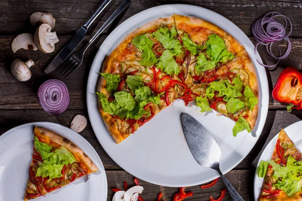 Pizza with pepper, salad, cheese, mushrooms, zucchini and onions in a plate on a wood table with cutlery and a slice of pizza on another plate