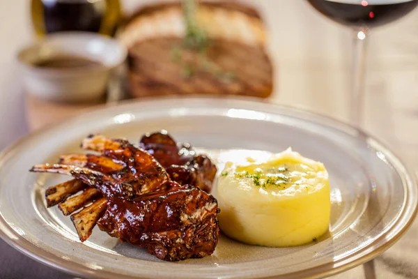 Grilled rack of lamb with barbecue sauce. Next to it is mashed potatoes with butter and dill. The food is on a round ceramic plate with sauce smeared on the plate. The plate is on a beige tablecloth, next to it is a wooden board with grilled toast, t