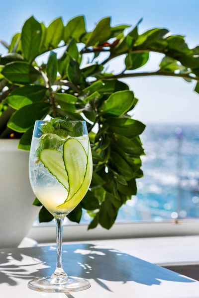 A cocktail with ice, cucumbers and mint in a glass that stands on the table against a background of blue sky and water