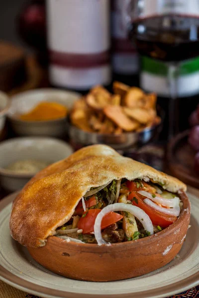 Domates, kabak, soğan, patlıcan ve hamur kapağıyla pişirilmiş havuç. Sebzeler odun ateşinde pişiriliyor. Tencere seramik tabakta. Yakınlarda, Gürcistan baharatlı seramik kaseler, bir bardak kırmızı şarap ve metal kâse wi var.