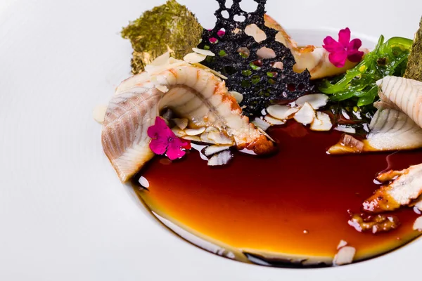 Tütsülenmiş yılan balığı ve balzamik soslu panna cotta, üstünde chuka salatası, badem gevreği, yenilebilir taç yaprakları, yaldızlı Norveç yosunu. Yemek derin beyaz bir tabakta. Tabak beyaz bir masa örtüsünün üzerinde, çatal bıçak takımı yanında, gri bir mineral camı var.