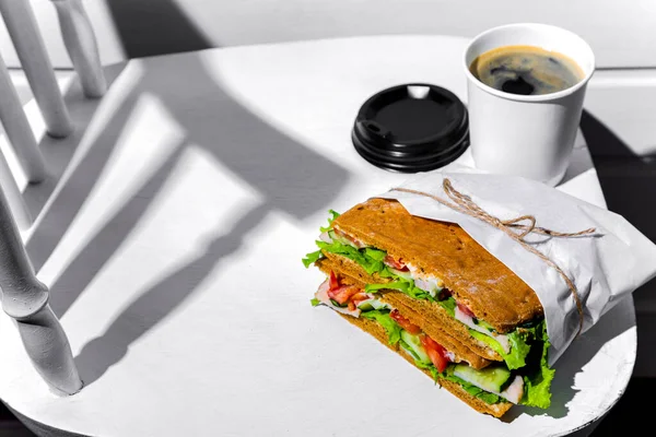 Sandwich in white paper with ham, lettuce, tomatoes, cucumbers and sauce with a paper cup of cappuccino and a black lid standing on a white chair against a background of a white wooden wall