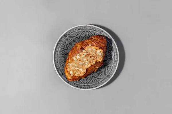Airy croissant with almonds and almond paste. The croissant lies on a gray, round, patterned ceramic plate. The plate stands on a gray paper background.