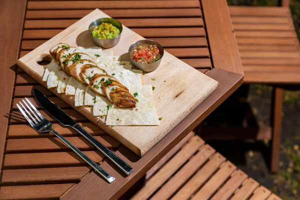 Pieces of meat with parsley, pita bread and two types of sauce in sauce bowls on a wooden board that lies on the table with cutlery