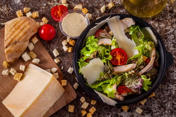 Salad with meat, tomatoes, lettuce, cheese, endive and sauce in a plate on a stone background with a piece of cheese, croutons, bread and olive oil