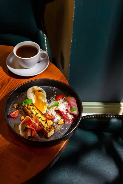 A slice of cheesecake with thyme, prosciutto, parmesan, basil, tomatoes, garlic and a boiled egg in a plate on the table with a cup of coffee