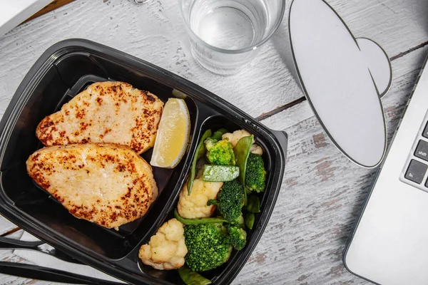 Fried cutlet with lemon, broccoli in a plastic container with a glass of water, and a laptop standing on a white wooden table