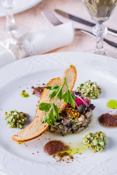 Herring tartar with cilantro, Dijon mustard, basil microgreen, cilantro branch and a spoonful of red horseradish. On top is a rye bread chip. Nearby is a finely chopped cucumber with Dijon mustard. The food is in a round, white plate. The plate is on