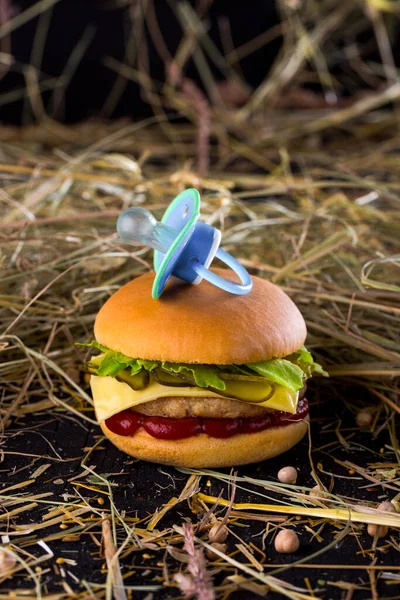 Mini burger with bun and baby pacifier on top, cutlet, pickled cucumbers and ketchup on the table with chickpeas and sprinkled hay on a black background. Vertical orientation