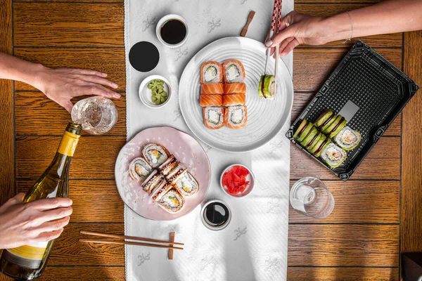 Two people eat roles and drink champagne. Rolls with eel, salmon, sesame seeds, Philadelphia cheese, avocado, shrimp, cucumber, in plates and plastic containers with soy sauce in a bowl, pickled ginger and wasabi standing on the table