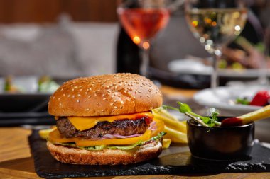 Hamburger with cutlet, cheese, tomatoes, onion, frisse salad, sesame and ketchup, sauce in a saucepan and pea sprouts. Everything stands on a stone slab against the background of glasses of wine