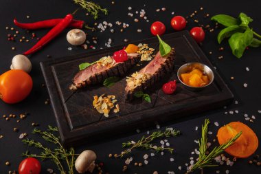 Pork steak with almond slices, cherries, peaches, mint on a wooden board with mushrooms, peas, red chili peppers, salt, tomatoes and basil on a black background
