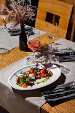 Salad with strawberries, figs, pea sprouts, mozzarella, flower petals, pesto sauce, mung bean salad, beans and pumpkin seeds in a plate on the table with glasses of wine, cutlery, a vase and dried flowers, a tablecloth and a napkin on the background