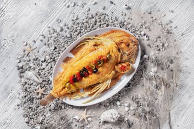 Fried dorado with tomatoes, lemon and olives in a plate standing on a white wooden table with seashells, stones and leaves