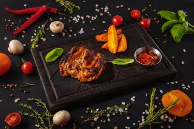 Grilled meat steak in sauce with fried potatoes, basil and ketchup on a wooden board with mushrooms, peas, red chili peppers, salt, tomatoes and basil on a black background