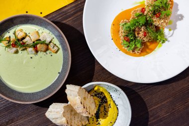 Breaded cutlets with onions, parsley, sauce and pepper in the first plate, hummus with sesame seeds and bread in the second plate and cream soup with croutons in the third plate standing on the table