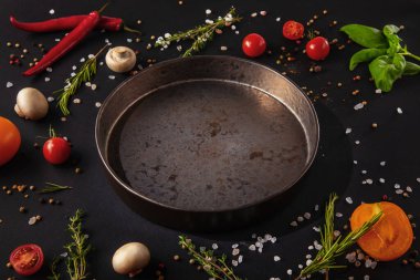 Iron plate stands on a dark background with mushrooms, peppercorns, red chili peppers, salt, tomatoes and basil on a black background