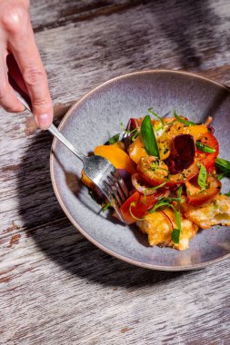 Domatesli salata, bezelye filizi, çiçek yaprakları, ekmek ve fesleğen bir tabakta ahşap bir masada eller ve çatal bıçaklarla.