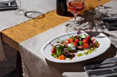 Salad with strawberries, figs, pea sprouts, mozzarella, flower petals, pesto sauce, mung bean salad, beans and pumpkin seeds in a plate on the table with glasses of wine, cutlery, a vase and dried flowers, a tablecloth and a napkin on the background