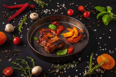 Grilled meat steak in sauce with fried potatoes, basil and ketchup on an iron plate with mushrooms, peas, red chili peppers, salt, tomatoes and basil on a black background