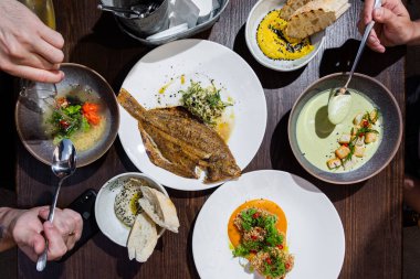 İnsanlar tabakları masada yerler. Birinci tabakta soğanlı pirzola, maydanoz, sos ve biber, iki tabakta susamlı humus ve ekmek, dördüncü tabakta kremalı ekmek ve somon, maydanoz ve salata ile çorba.